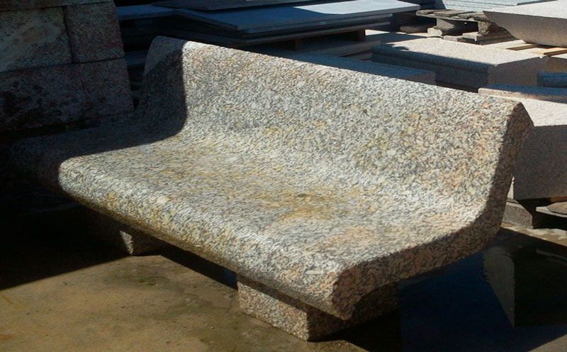 Un banco de piedra moteada de color beige y gris, con respaldo curvo, situado al aire libre en un taller o patio de piedra.