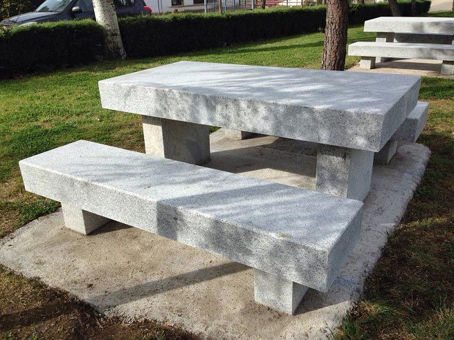 Una mesa de picnic de piedra con dos bancos a juego se encuentra sobre una base de hormigón en una zona de césped con árboles.