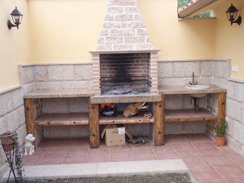 Una zona de barbacoa al aire libre revestida de piedra, con una parrilla central de ladrillo, encimeras de piedra, un pequeño fregadero y soportes de madera.
