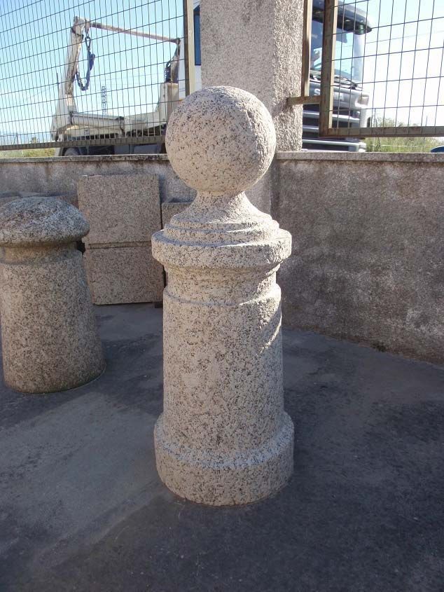 Un bolardo de piedra de color claro con la parte superior esférica, colocado sobre una superficie de hormigón cerca de una valla.
