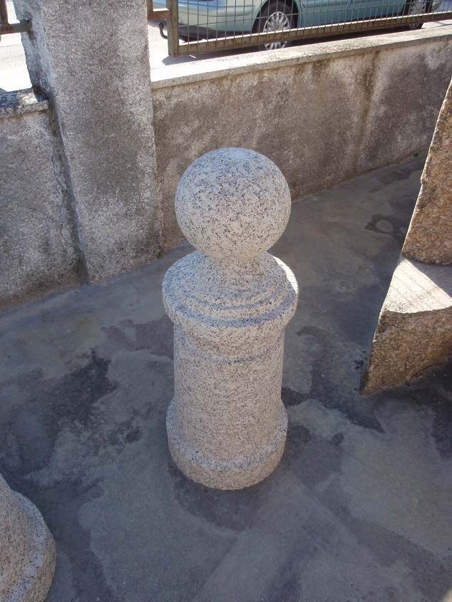 Un bolardo de piedra gris claro moteada con la parte superior esférica, situado sobre una superficie pavimentada cerca de un muro bajo.