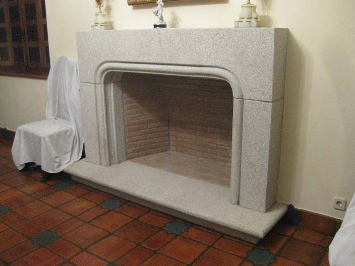 Una chimenea de piedra gris claro con una abertura arqueada, situada contra una pared lisa sobre un suelo de baldosas.