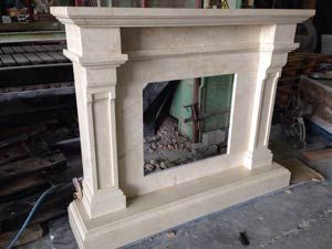 Una repisa de chimenea de piedra color crema ubicada en un taller, con columnas clásicas y una abertura rectangular y limpia.