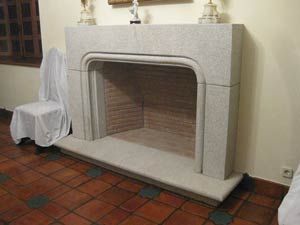 En una habitación con suelo de baldosas de terracota y paredes blancas, se alza una repisa de chimenea de piedra de color claro.