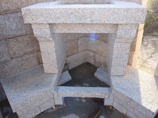 Una chimenea de esquina construida con bloques de piedra moteada de color claro, con una abertura central y bancos laterales.