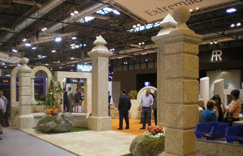 Pilares de piedra expuestos en un stand de una feria comercial, con visitantes de pie cerca de arcos de piedra y jardines decorativos.