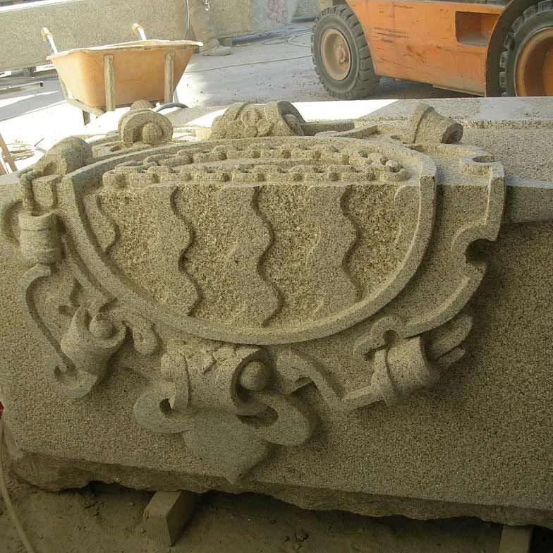 Un bloque de granito con una cresta decorativa finamente tallada, con líneas onduladas y motivos ornamentales, en un taller industrial.