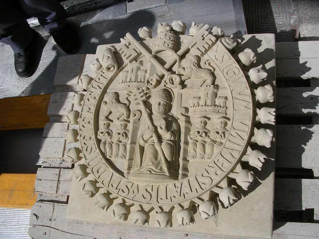 Un sello en relieve de piedra que representa una figura sosteniendo un bastón, rodeada de figuras más pequeñas y texto, que descansa sobre una plataforma de madera.