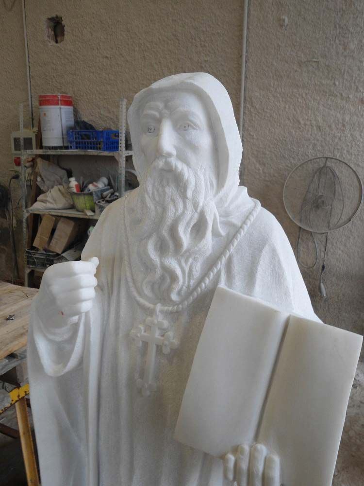 En un taller se encuentra una estatua de mármol blanco que representa una figura vestida con una túnica y barba larga, sosteniendo un libro abierto y portando una cruz.