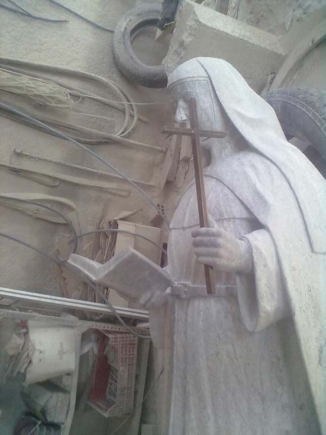 Una estatua de piedra blanca que representa a una figura religiosa con hábito, sosteniendo una cruz de madera y un libro abierto, situada en un taller desordenado.