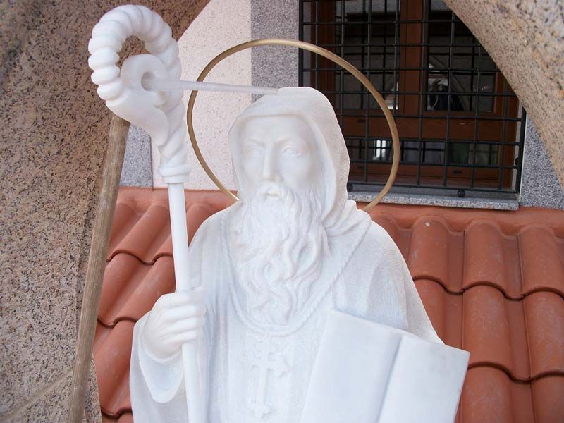 Estatua de mármol blanco de un santo barbudo con aureola, que sostiene un báculo y un libro, situada frente a un tejado de tejas.