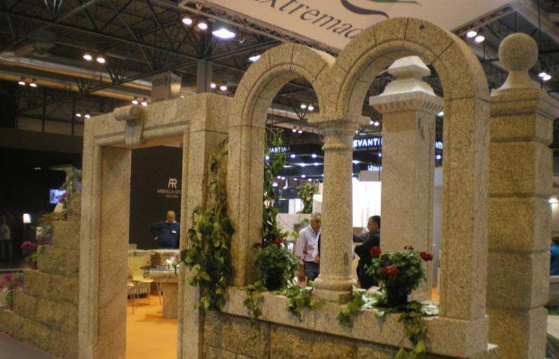 Una vitrina de piedra con una ventana arqueada y una puerta, decorada con vegetación, en un stand de una feria comercial interior.