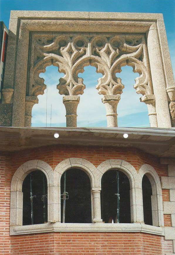 Una fotografía arquitectónica que muestra un marco de ventana de estilo gótico, ornamentado y tallado en piedra, sobre una pared de ladrillo con cuatro ventanas arqueadas.