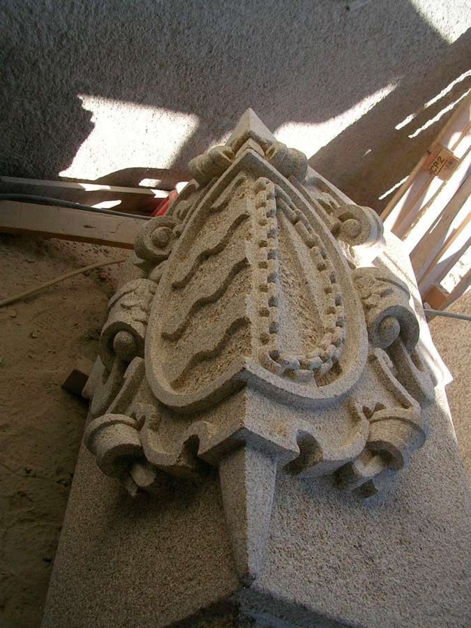 Una talla de piedra de color claro que presenta una cresta en forma de escudo con líneas onduladas y un borde de cadena, montada en una esquina.