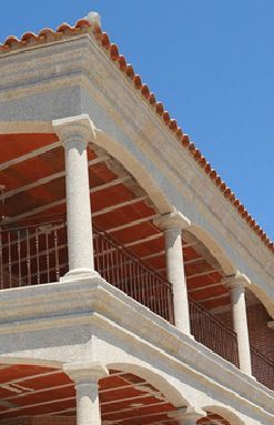 Vista desde un ángulo bajo de la esquina de un edificio con columnas de piedra que sostienen un balcón cubierto con techo de baldosas de terracota.