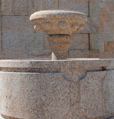Una fuente de piedra con una pila circular y un pedestal tallado, situada contra un muro de bloques de piedra.