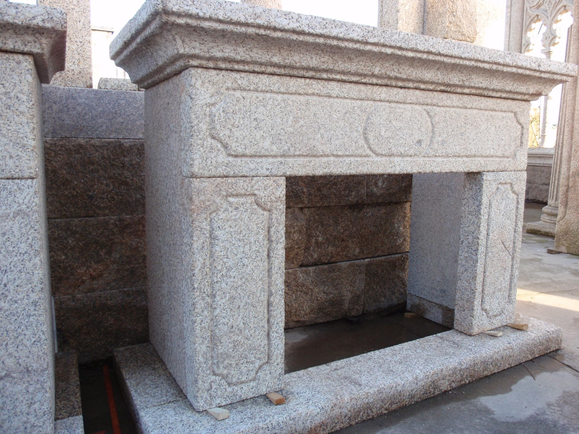 Una repisa de chimenea de granito gris claro con sencillos detalles rectangulares tallados, expuesta al aire libre.