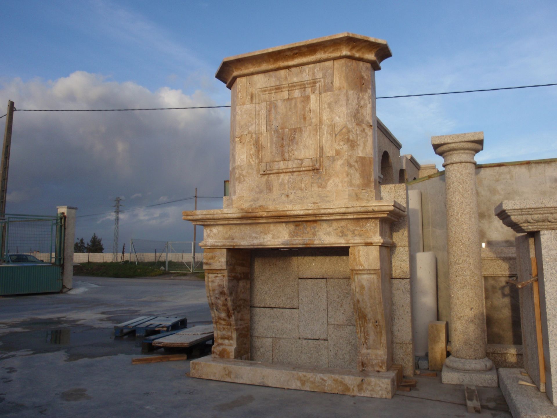 Una gran repisa de chimenea decorativa de piedra beige se encuentra al aire libre en un almacén, junto a una columna de piedra tallada.