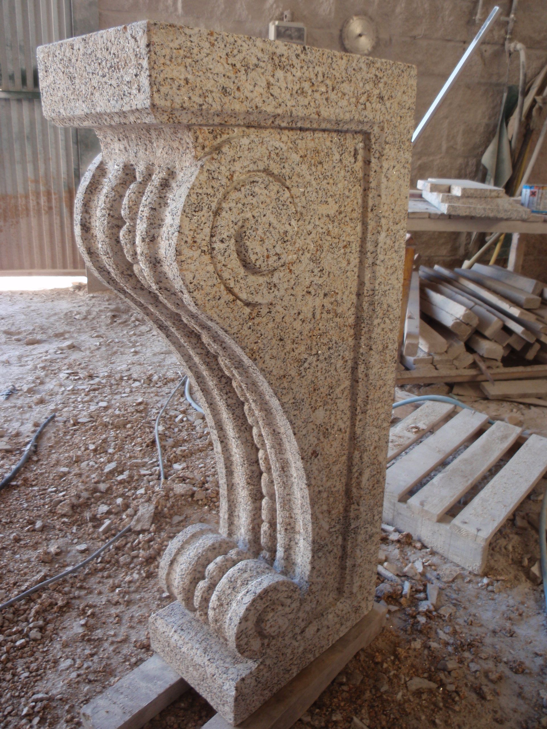 Una ménsula esculpida de granito beige con ornamentación de volutas se encuentra en un polvoriento taller de piedra.