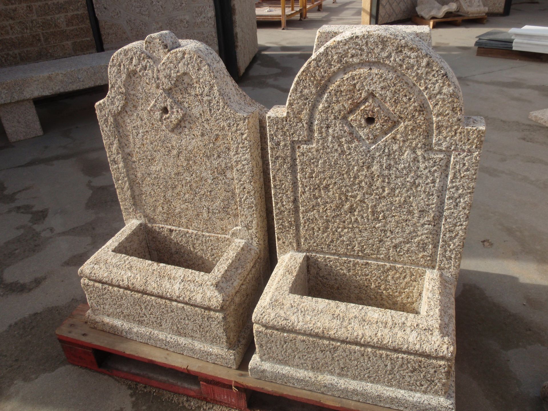 Dos fuentes de pared de piedra texturizada de color tostado, con sus respectivas pilas, se encuentran una al lado de la otra sobre un palé de madera en un patio de piedra al aire libre.