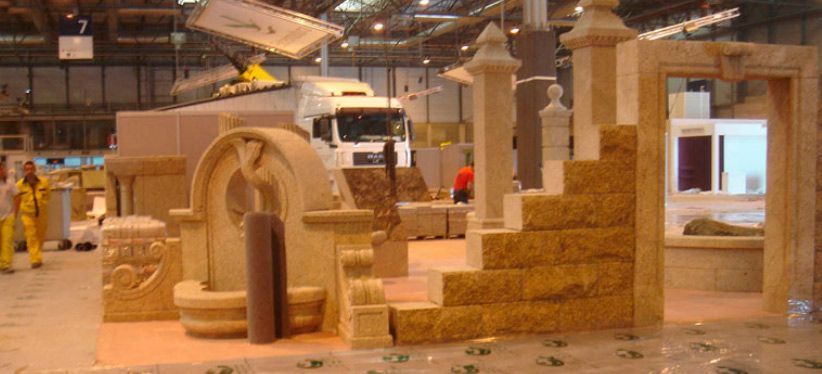 Una muestra de mampostería en piedra en una feria comercial bajo techo que incluye una fuente, escalones y un arco decorativo en la entrada.