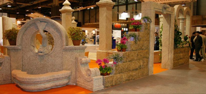 Fuente de piedra y exhibición arquitectónica con plantas en macetas en un stand de exposición interior.