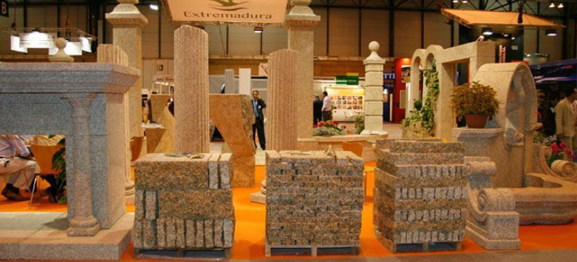 Un stand de feria comercial que exhibe diversos elementos arquitectónicos de piedra, como columnas, pedestales y bloques de piedra tallada.