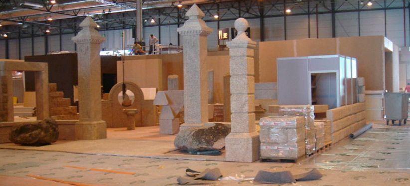 Una muestra de elementos arquitectónicos de piedra, incluyendo pilares y segmentos de muros, se exhibe en el interior de un gran pabellón de exposiciones comerciales.
