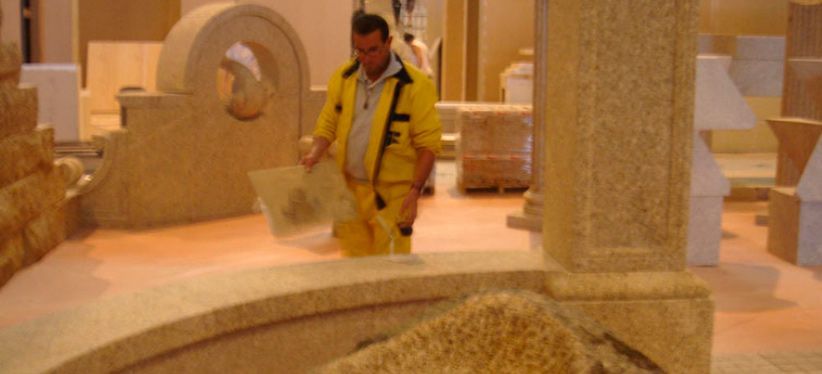 Un trabajador con una chaqueta amarilla inspecciona piezas arquitectónicas de piedra dentro de un taller o sala de exposiciones.