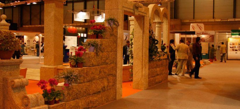 Un stand de feria comercial con pilares de piedra decorativos y arquitectura arqueada, adornado con macetas de flores rojas.