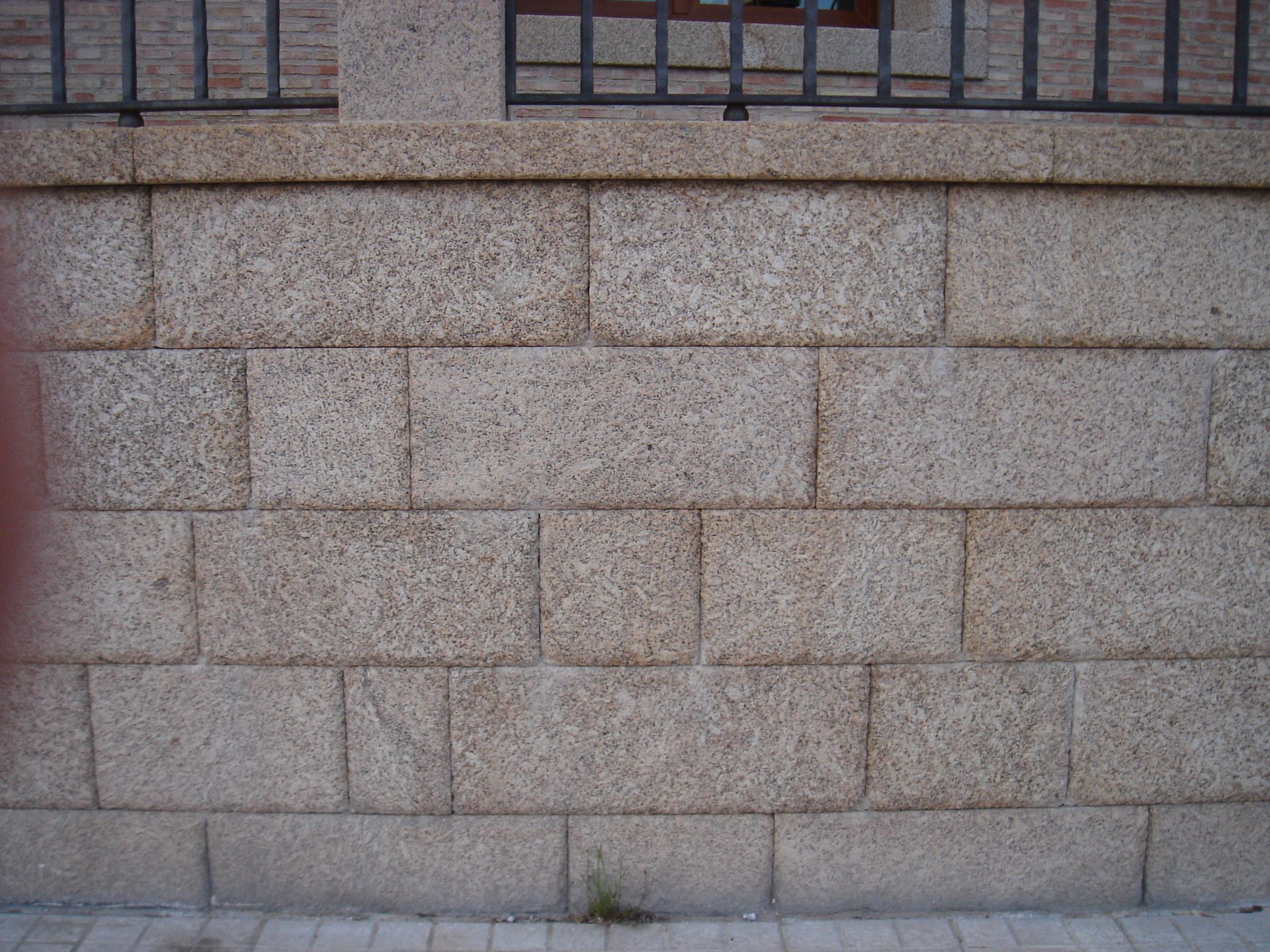 Un muro hecho de bloques rectangulares de granito de color marrón claro se encuentra debajo de una valla metálica negra.