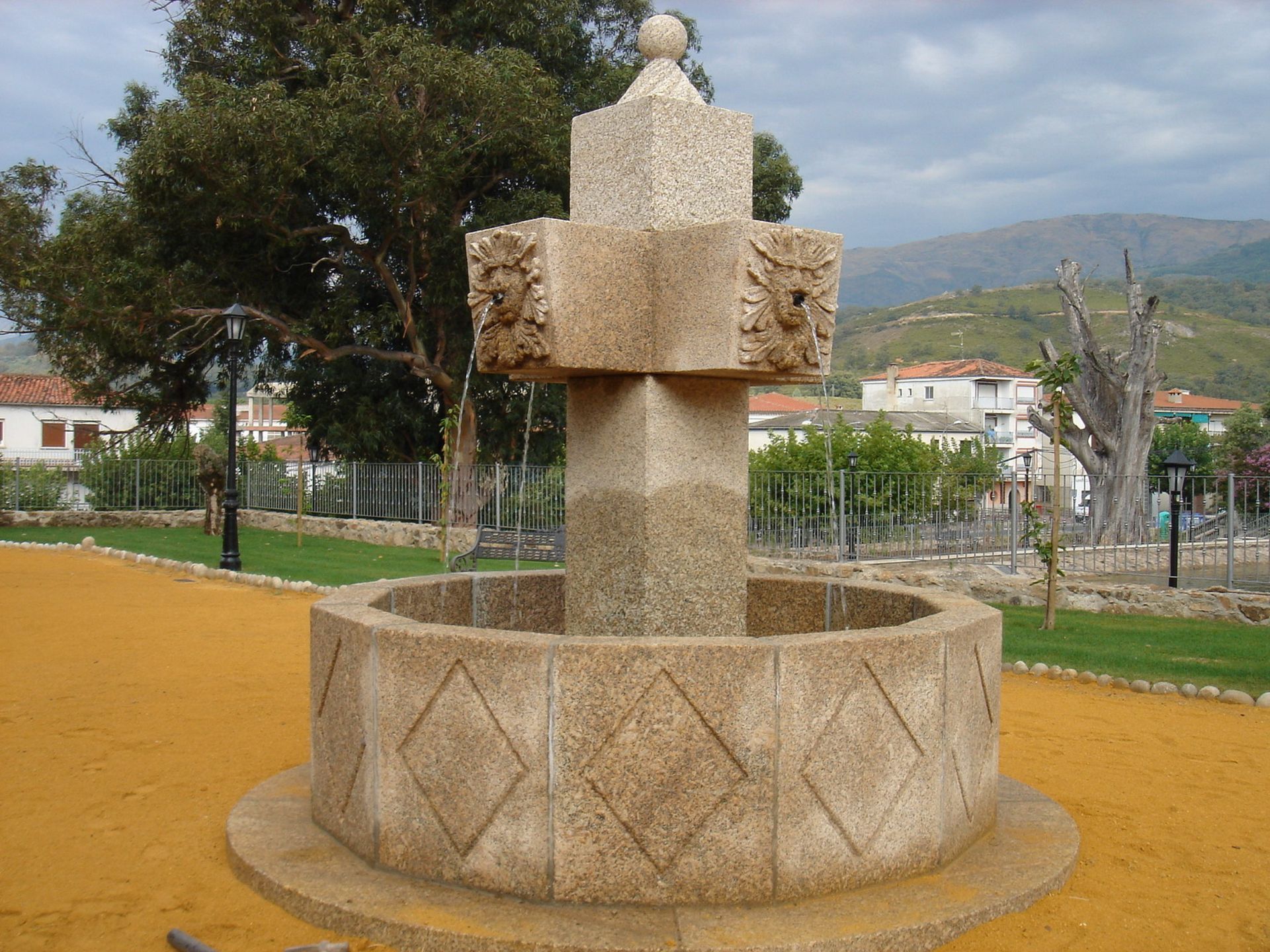 Una fuente de piedra con un pilar tallado y una pila central, situada en un parque con árboles y colinas al fondo.