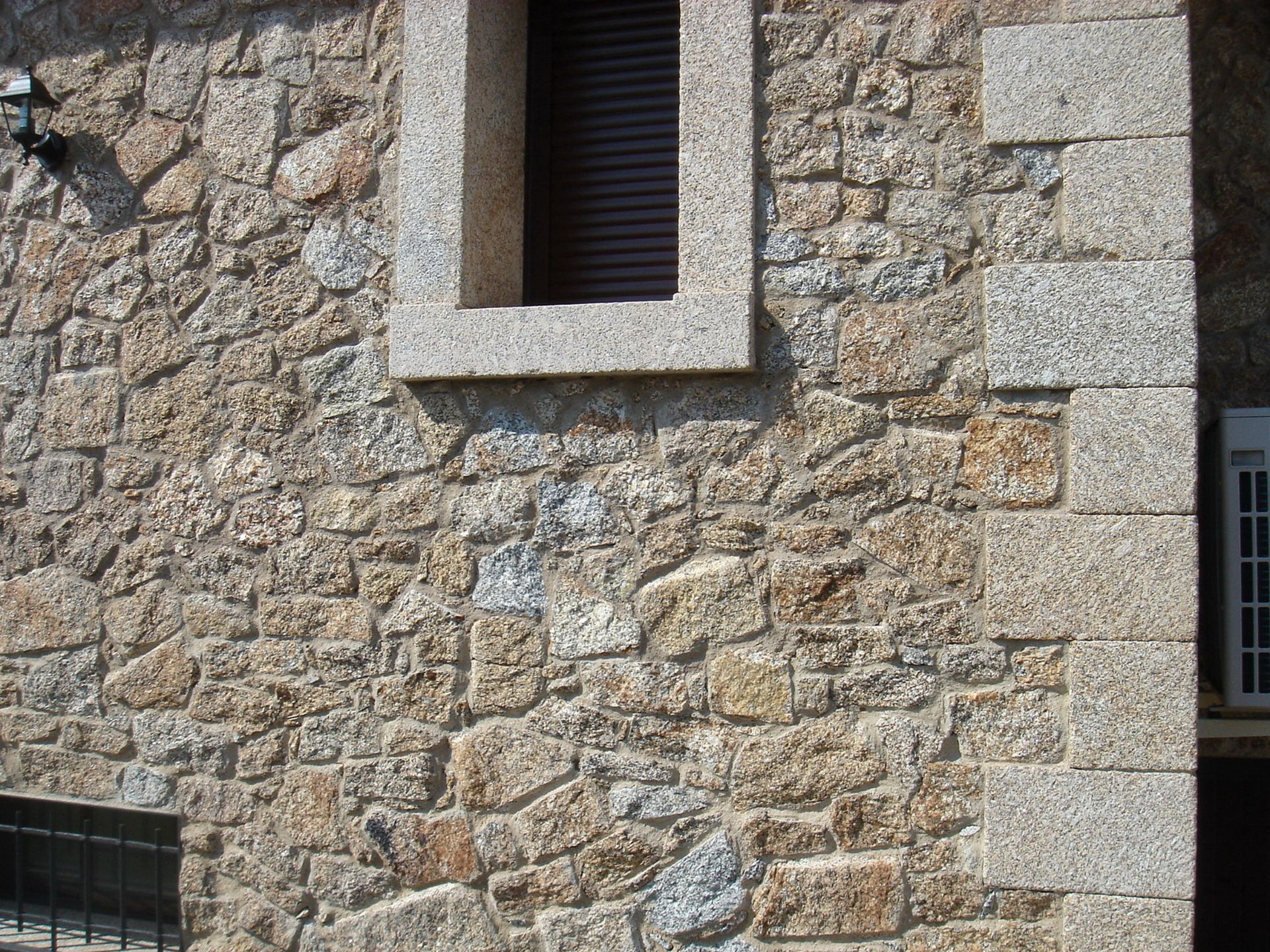 Muro de piedra texturizada con una ventana de contraventanas oscuras y una esquina de piedra enmarcada, bañado por la luz del sol.