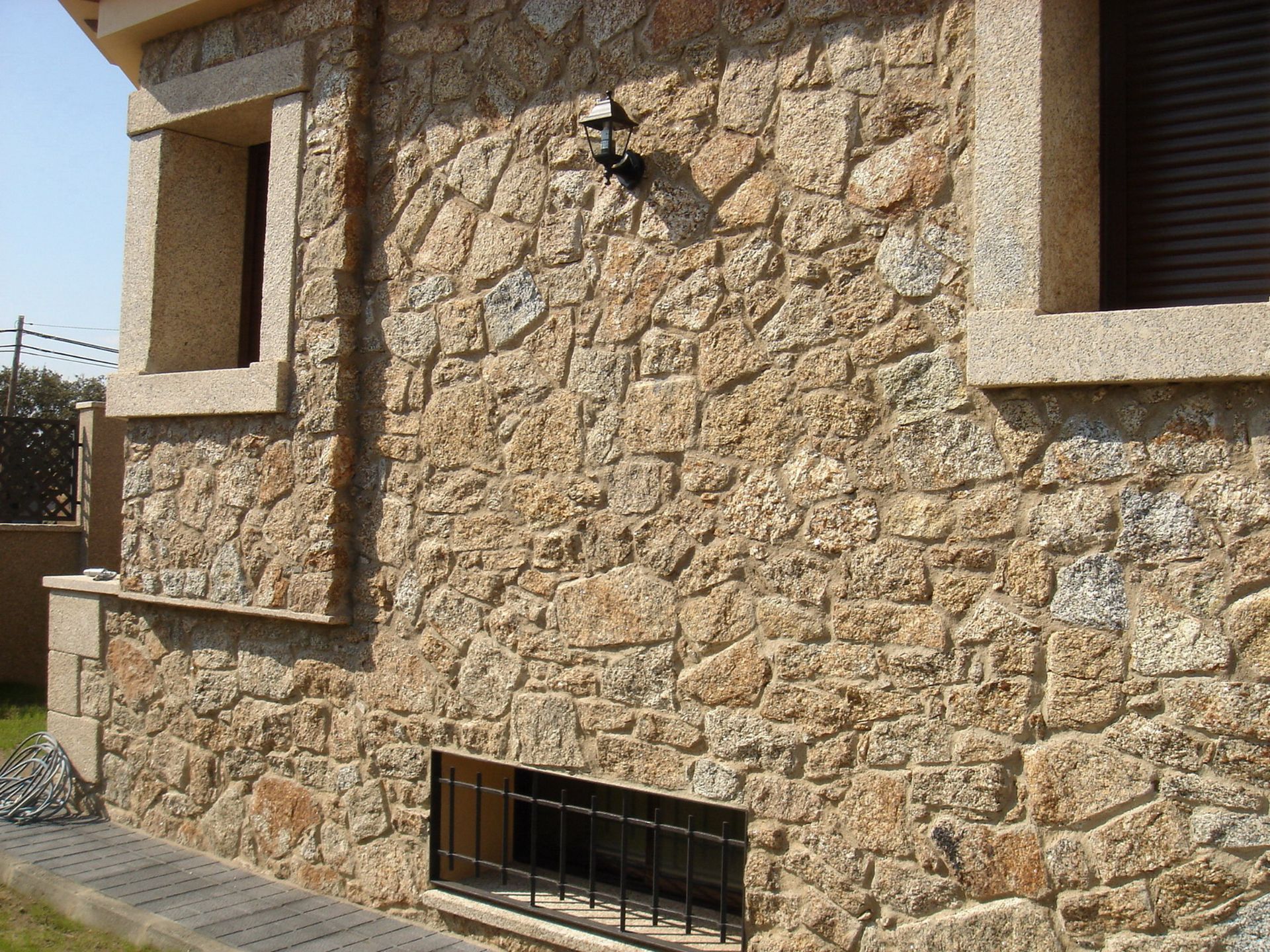 Una pared exterior de una casa hecha de piedra natural texturizada de color tostado, con una ventana en el sótano y una lámpara de pared.