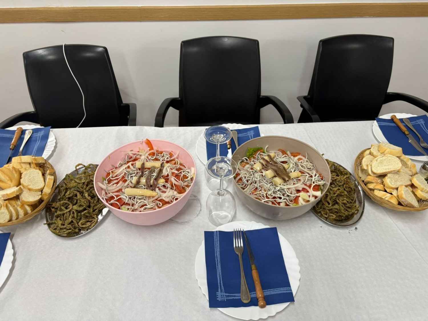 Mesa puesta con tres cuencos de comida, patatas fritas y servilletas sobre un mantel blanco, con sillas vacías detrás.