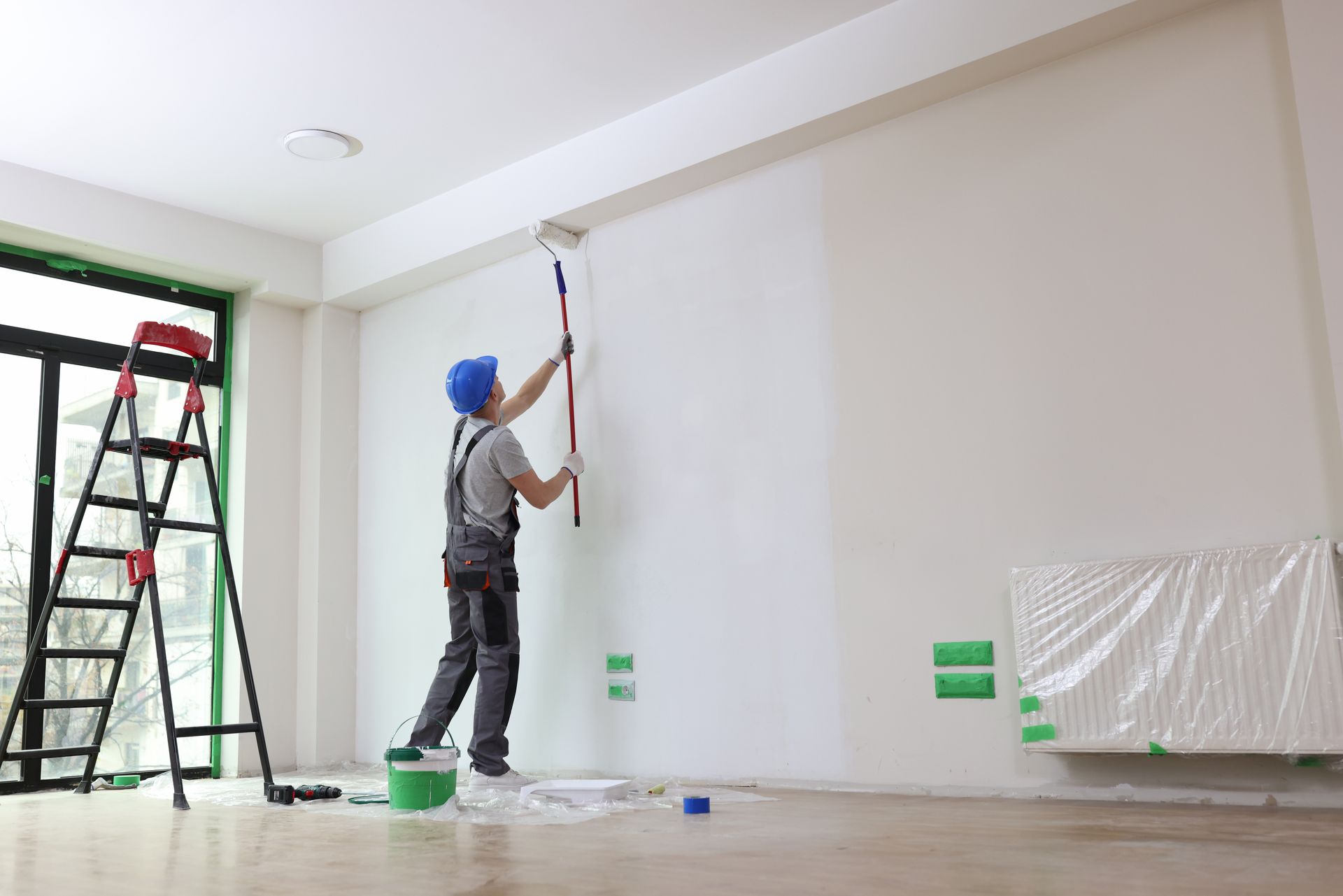 Un homme portant un chapeau bleu peint un mur blanc au rouleau. Décor intérieur, avec une échelle.