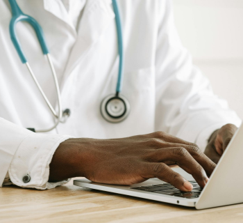 Médico de jaleco branco com estetoscópio digitando em um laptop.