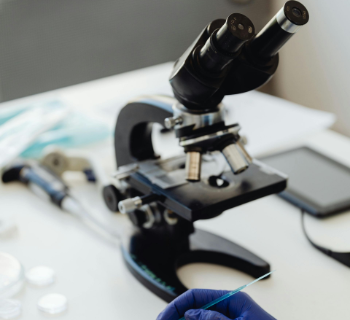 Cientista com luvas azuis utiliza um instrumento para examinar um líquido verde em uma placa de Petri com um microscópio