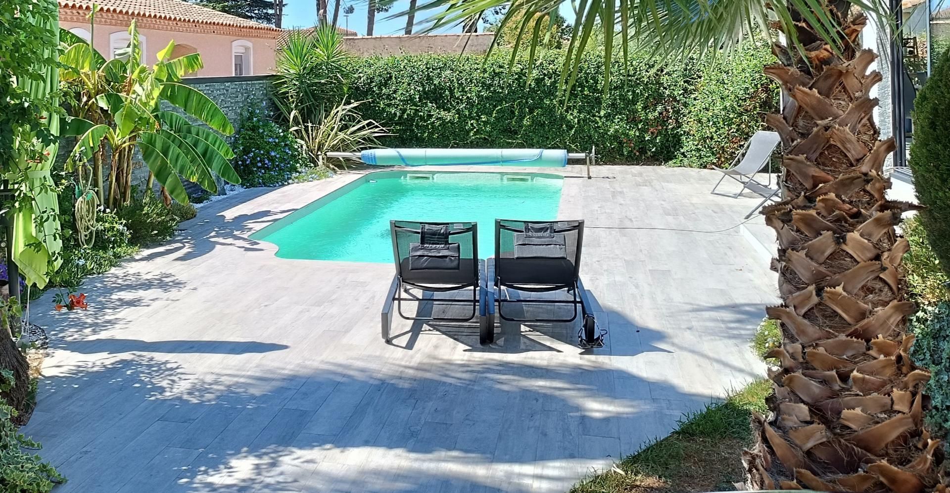 Piscine dans le jardin avec deux chaises longues sur une terrasse ensoleillée, encadrée de palmiers et de verdure.