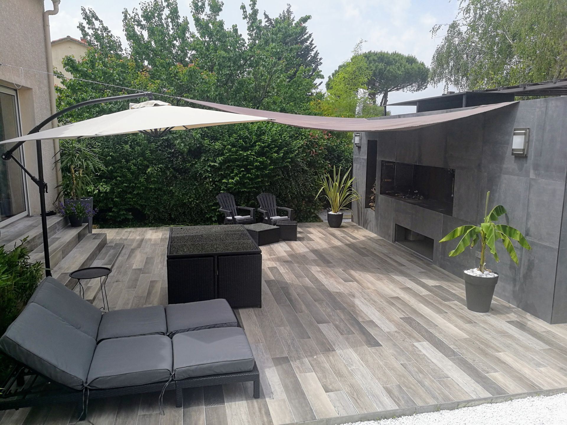 Terrasse moderne dans un jardin : chaises longues grises, parasol, ensemble repas et cuisine extérieure près d'une haie.