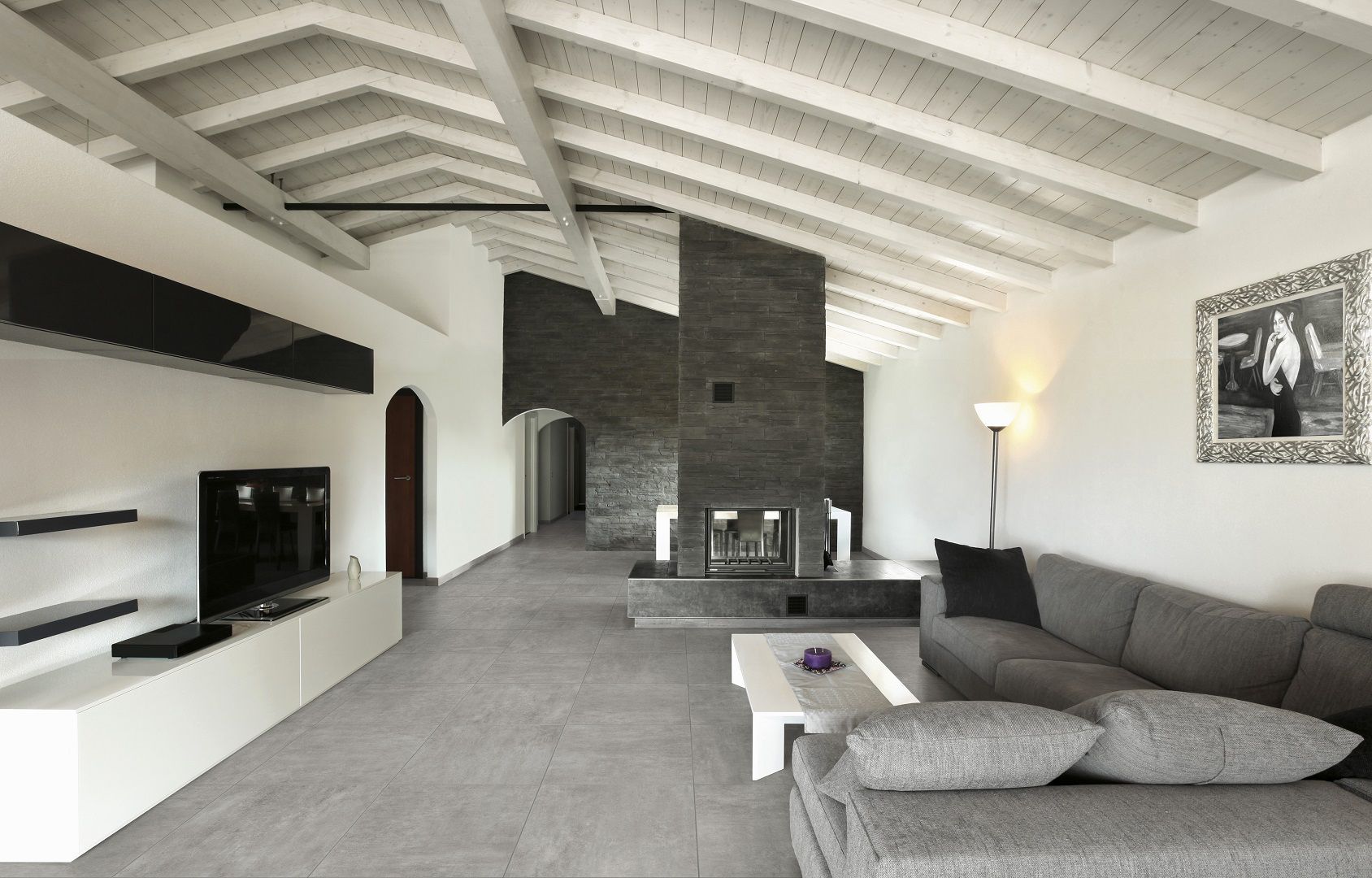 Salon moderne et lumineux avec canapé d'angle gris, plafond voûté, cheminée, télévision et table basse blanche