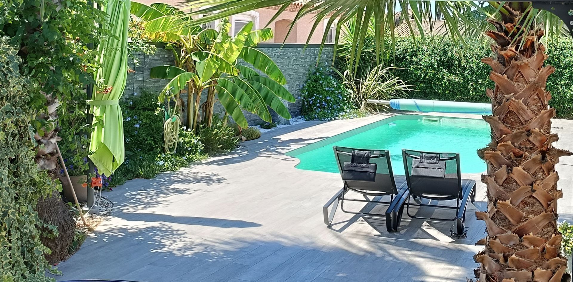 Terrasse ensoleillée avec piscine dans le jardin, deux chaises longues, palmiers et végétation luxuriante.
