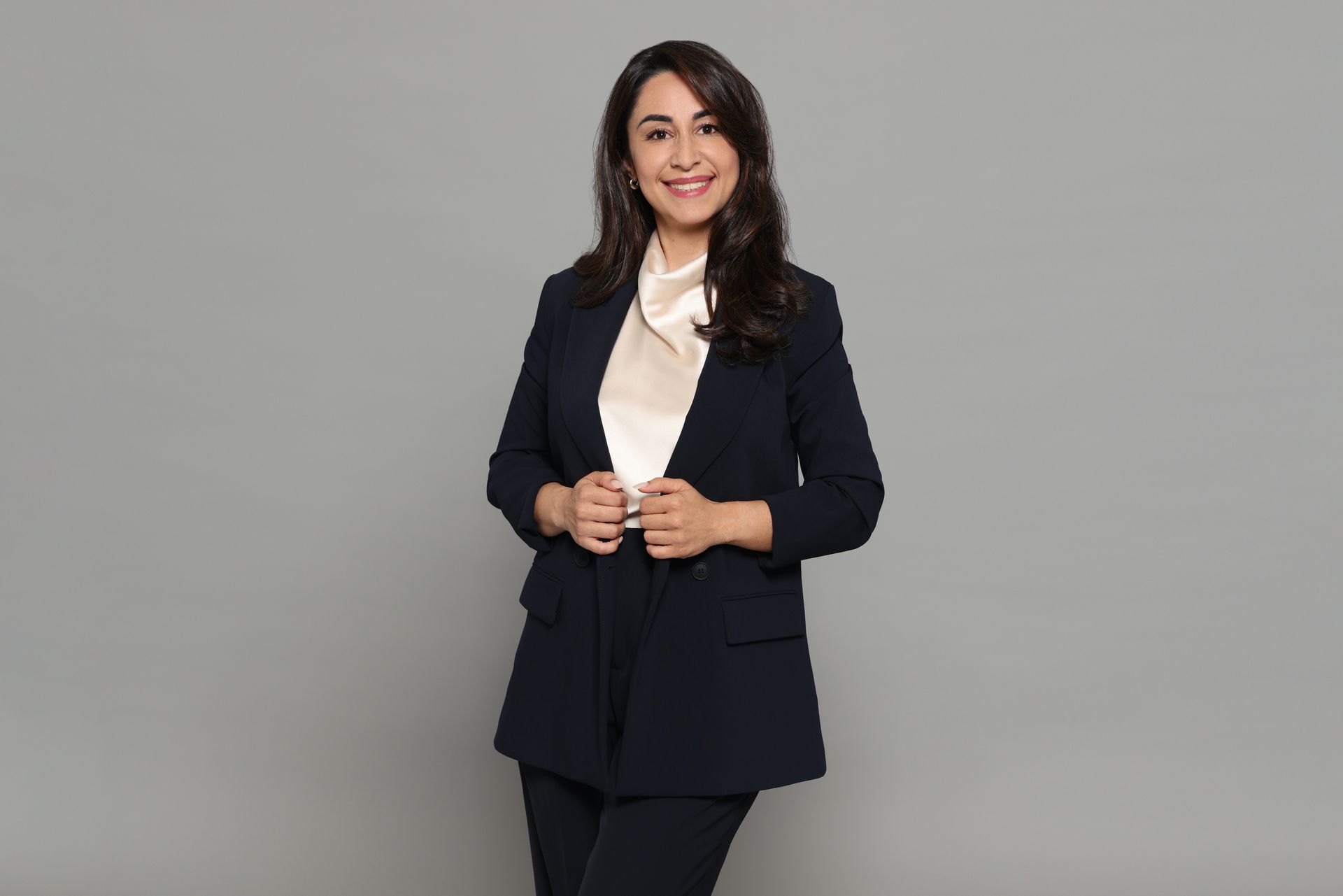 Mujer con traje oscuro de pie frente a un fondo gris, sonriendo a la cámara.
