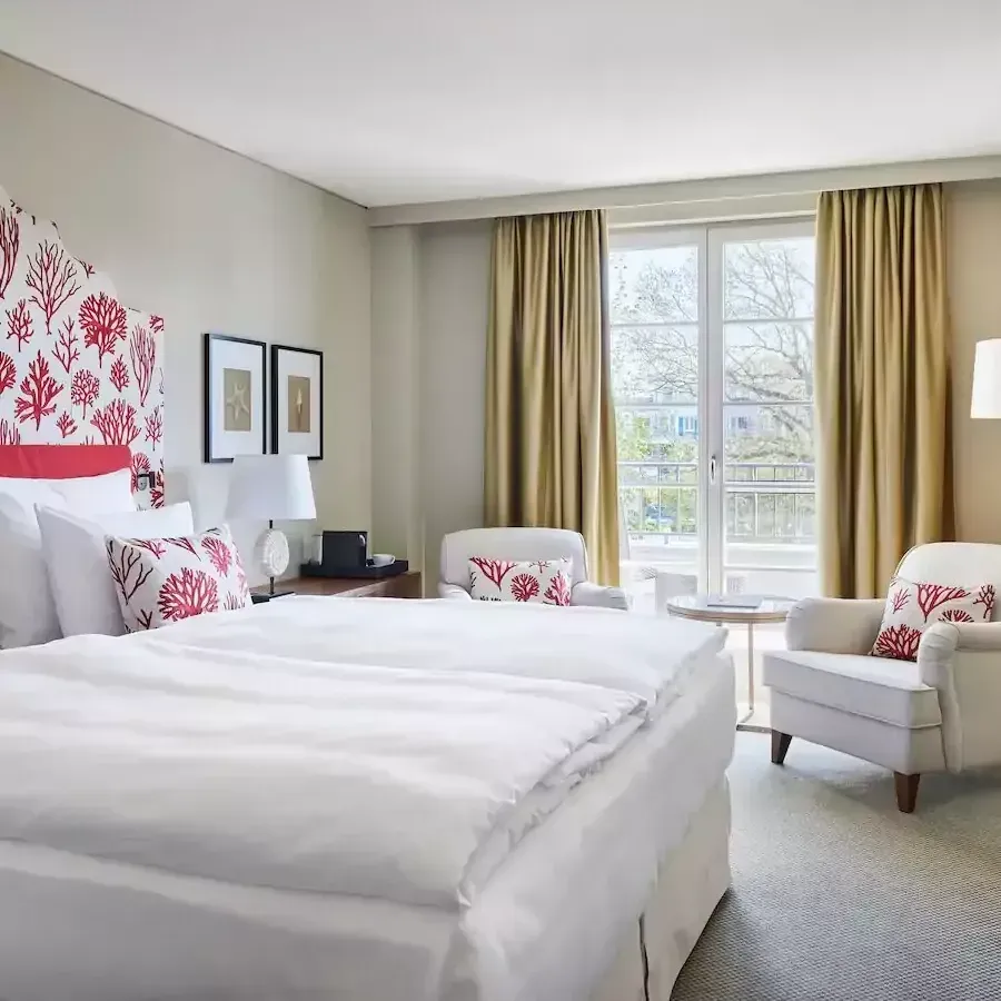 Schlafzimmer mit weißer Bettwäsche, korallenfarbenem Kopfteil und Kissen, zwei Sesseln und einer Balkontür.