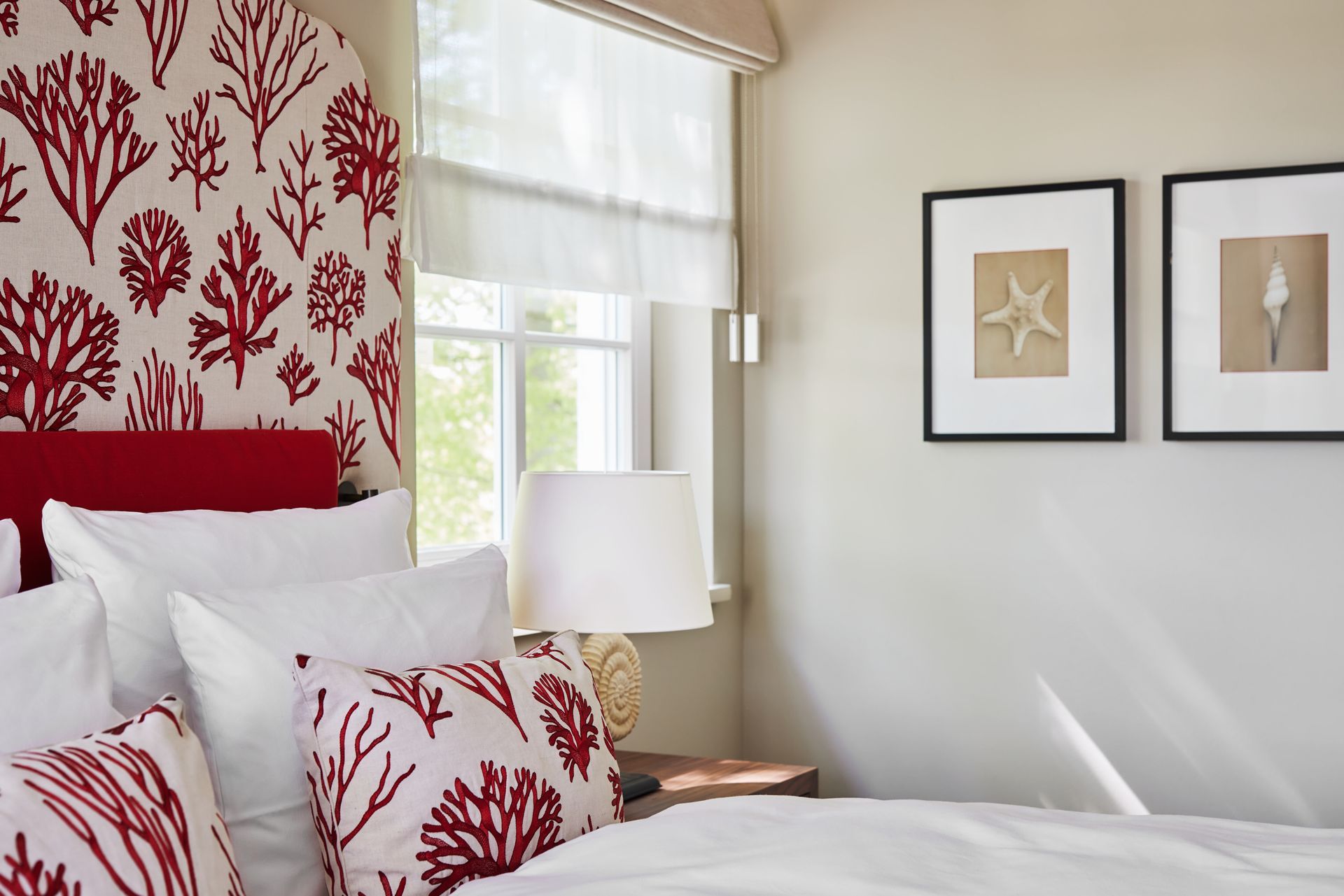 Schlafzimmer mit Kopfteil und Kissen mit rotem Korallenmuster, weißer Bettwäsche und gerahmter Muschelkunst.