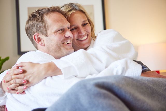 Ein Paar in weißen Bademänteln lächelt und umarmt sich im Hotelzimmer.