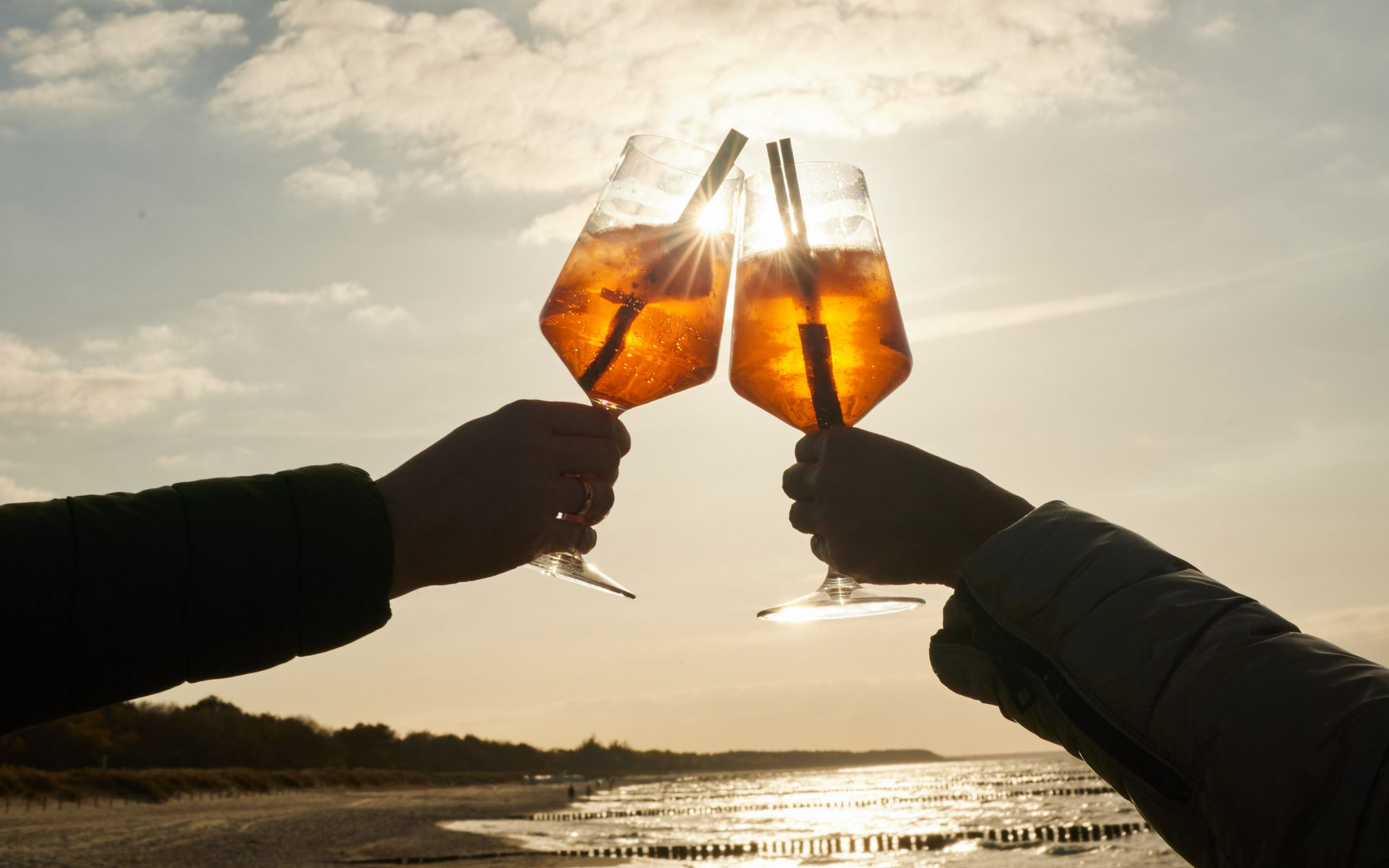 Zwei Hände klirren an bernsteinfarbenen Getränken vor einem Sonnenuntergangshimmel mit Wolken am Strand.