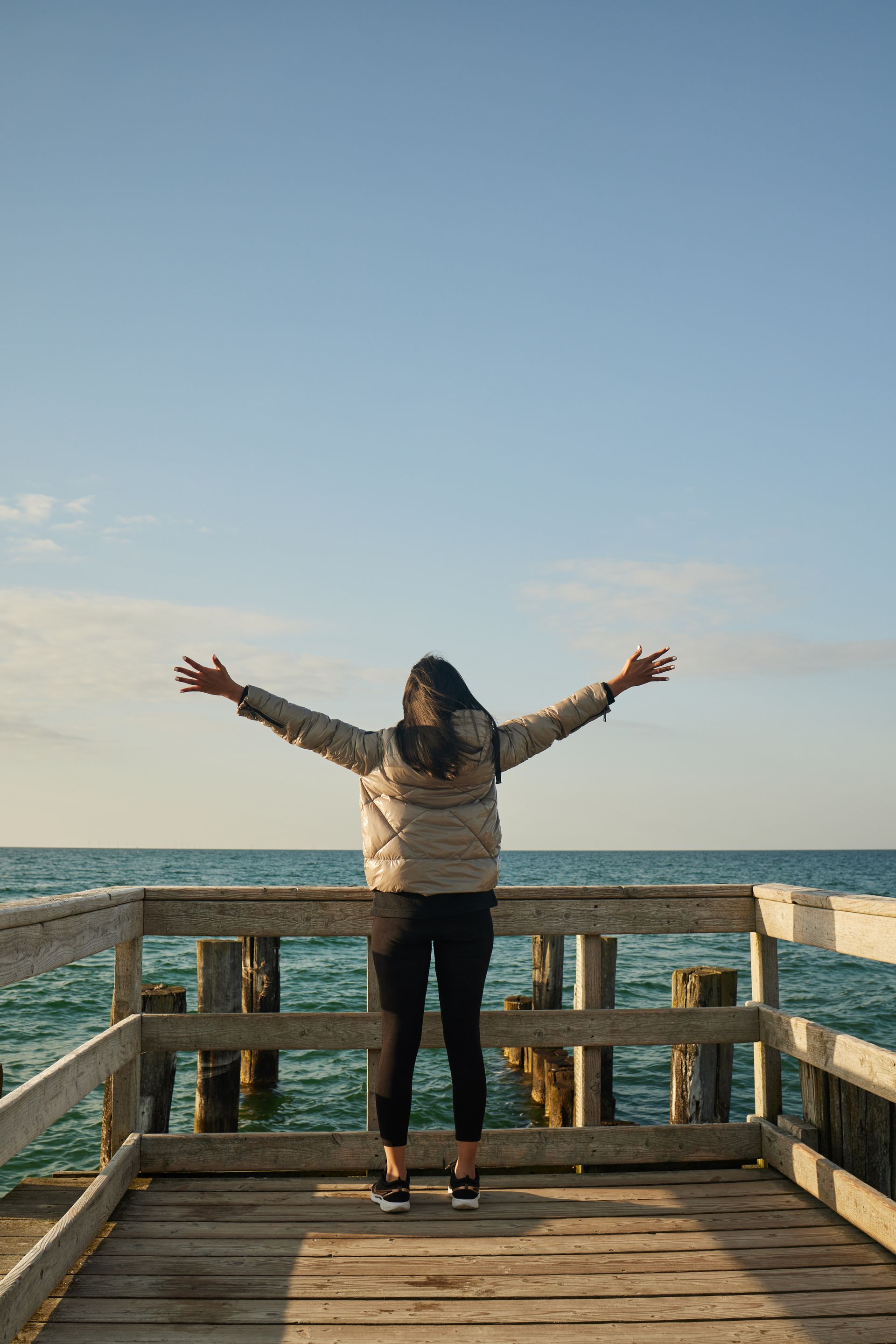 Person mit ausgestreckten Armen auf einem Holzsteg, mit Blick auf die Ostsee unter blauen Himmel.