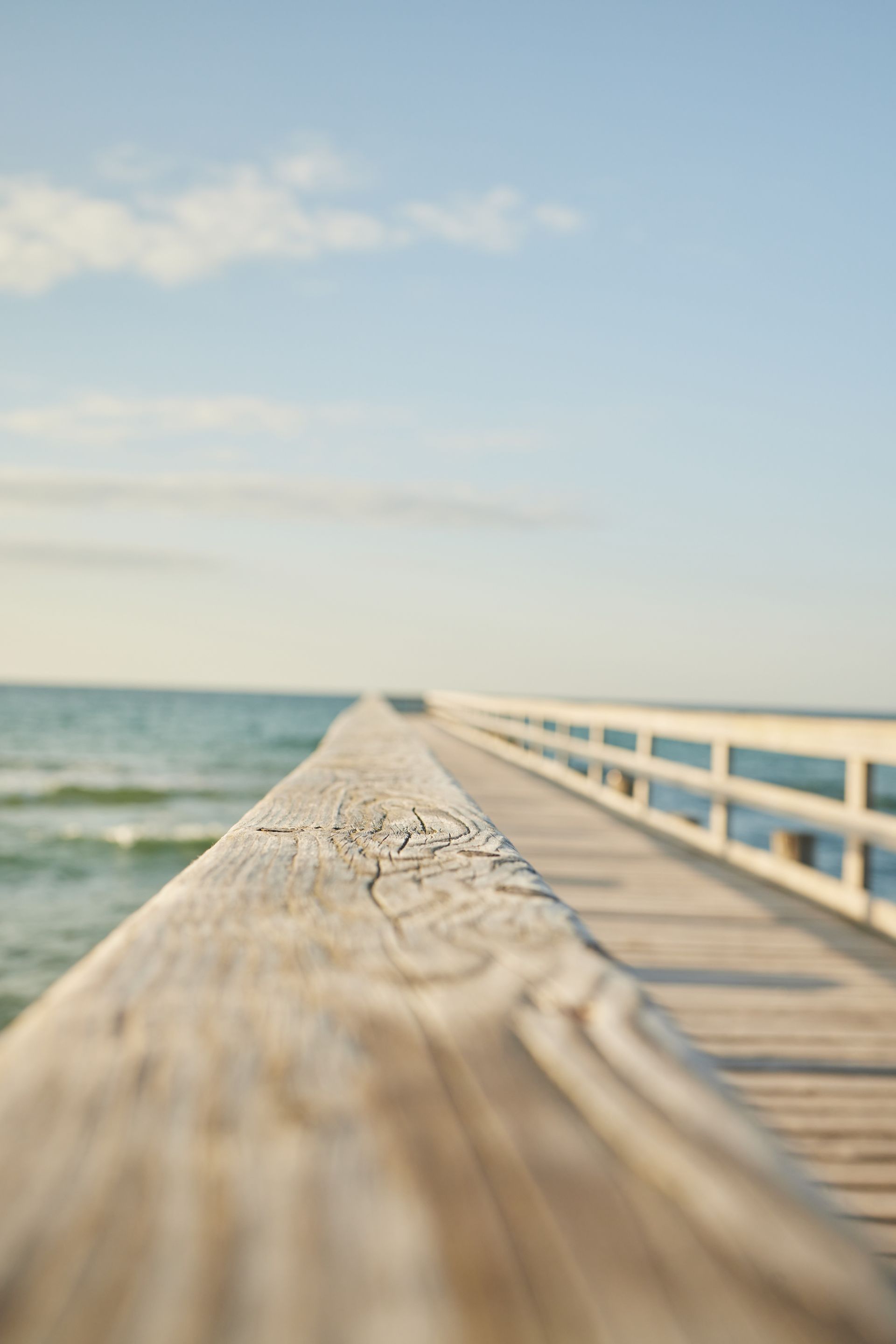 Holzsteg über dem Meer, blauer Himmel im Hintergrund, Geländer scharf im Fokus. Warmes, weiches Licht.
