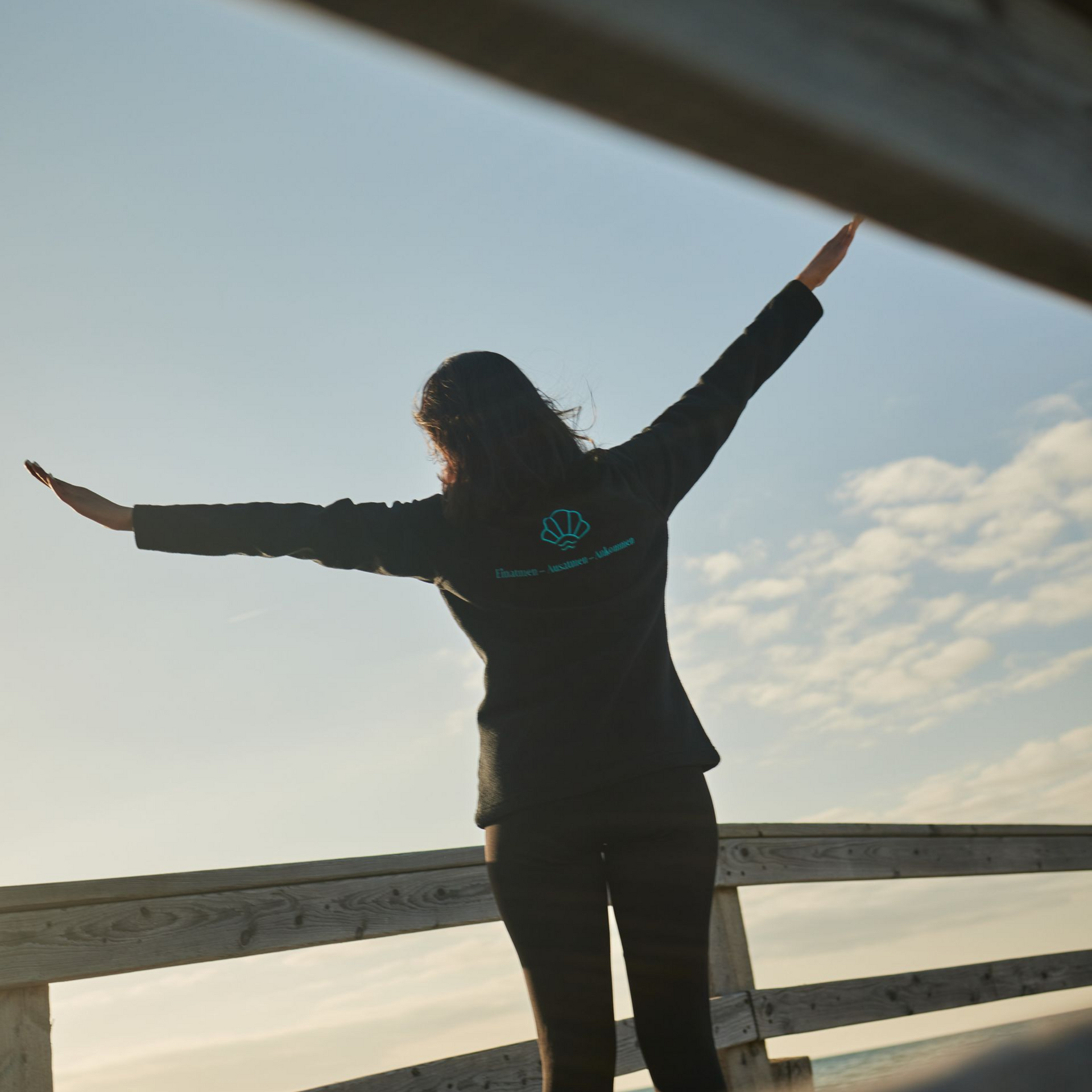 Person mit ausgestreckten Armen auf einem Holzsteg, mit dem Rücken zur Kamera, mit Blick auf das Meer.